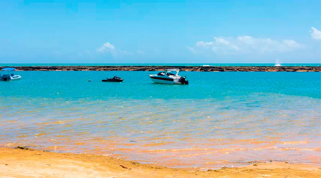 Lancha e jet ski ancorados no mar calmo da Praia de Barra de São Miguel. A longa barreira de recifes é visível no horizonte, separando as águas tranquilas da praia do mar aberto.