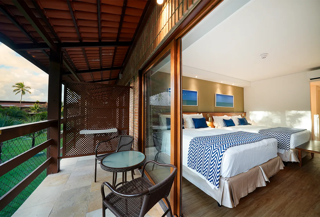 Varanda da acomodação Praia Jardim, com mesa, duas cadeiras e vista pro jardim do Salinas Maragogi All Inclusive Resort. Ao lado direito, a parte interna do quarto com camas queen size e quadros pendurados na parede.