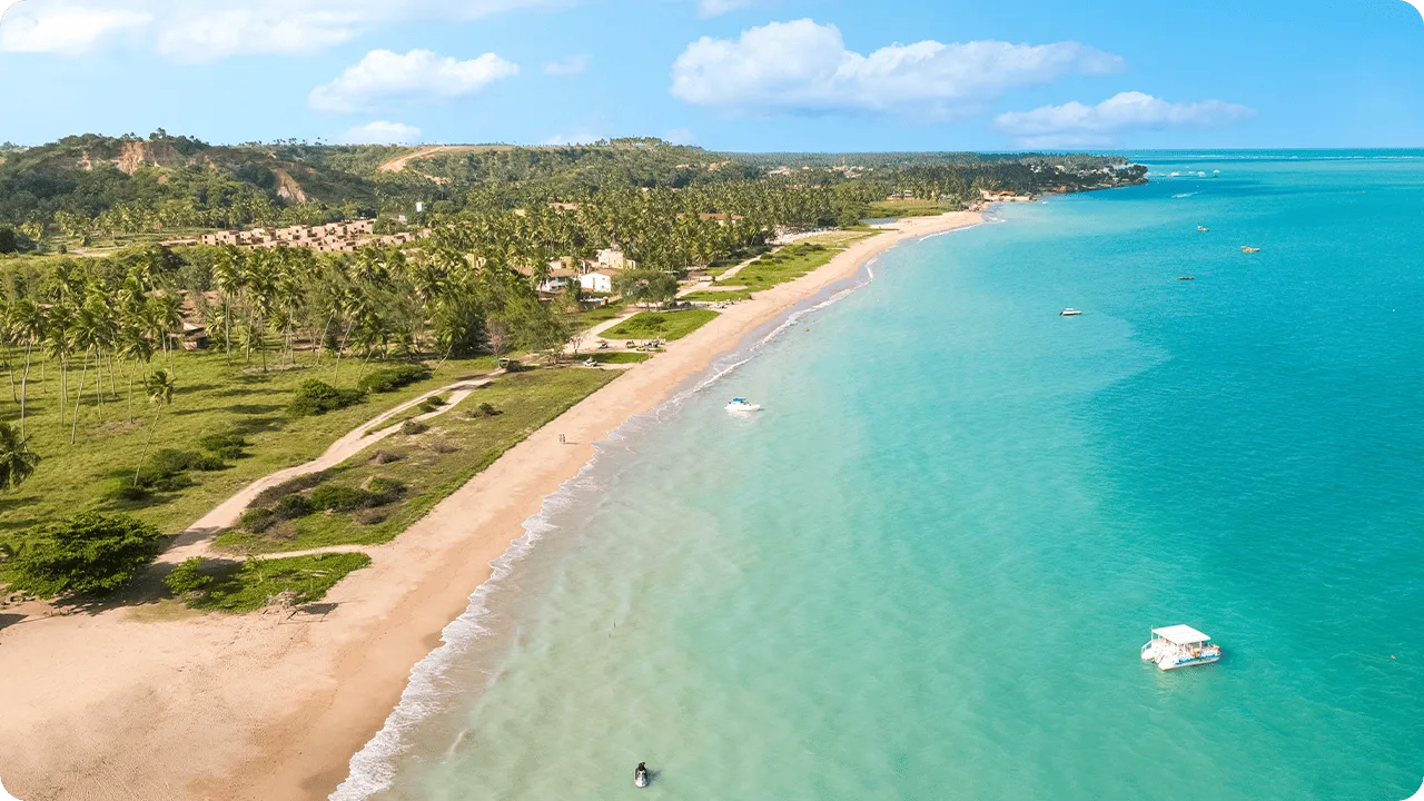 Vista aérea da Praia de Burgalhau com mar turquesa, faixa de areia clara, barcos espalhados, vegetação tropical e morros ao fundo
