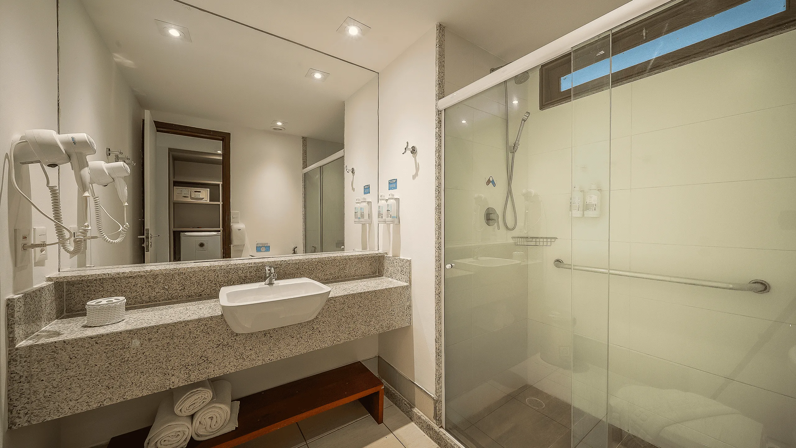 Banheiro do Apartamento Standard no Salinas Maceió com box de vidro, chuveiro, bancada ampla em granito com secador de cabelo pendurado na parede, e um espelho até o teto. 