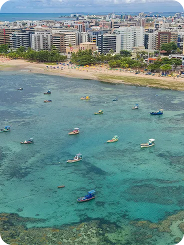 Vista aérea da praia de Jatiúca com águas rasas azul-turquesa, recifes de corais, barcos coloridos, guardas-sol coloridos na areia e prédios à beira-mar.