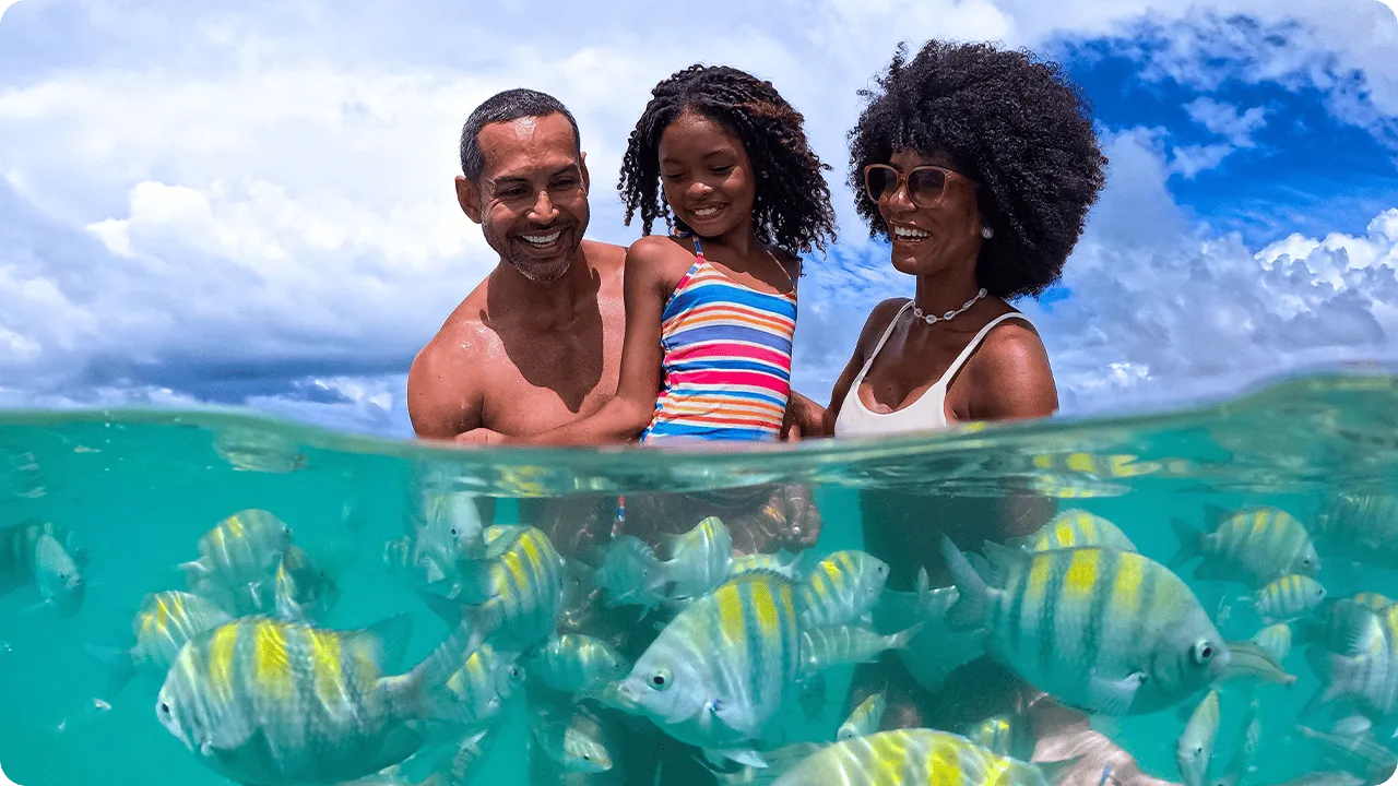 Família sorrindo em mergulho em dia ensolarado na Praia de Pajuçara com peixinhos coloridos nadando em águas turquesa