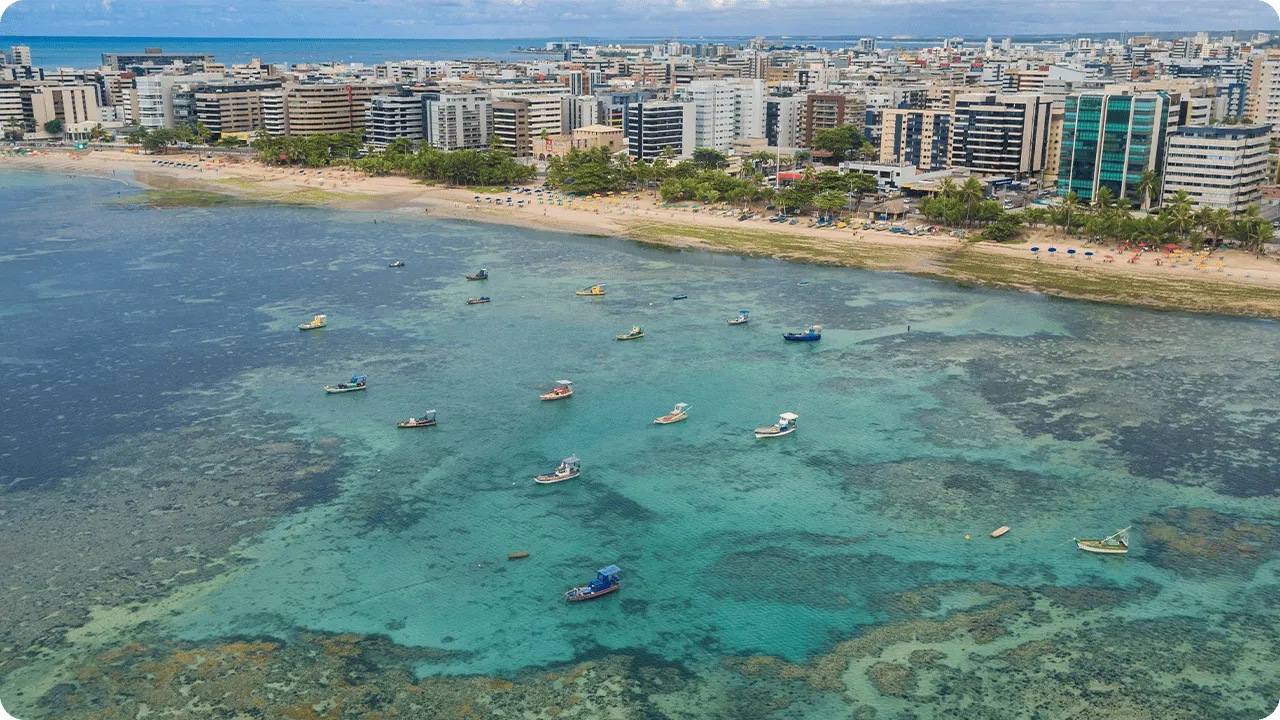 Vista aérea da praia de Jatiúca com águas rasas azul-turquesa, recifes de corais, barcos coloridos, guardas-sol coloridos na areia e prédios à beira-mar.