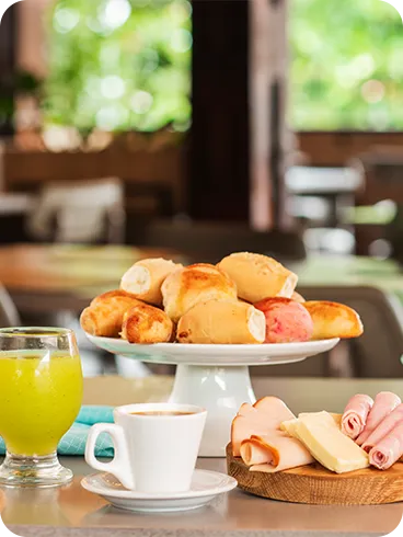 Mesa com pães, frios, queijos, bolos, geleia, café e sucos coloridos em ambiente iluminado