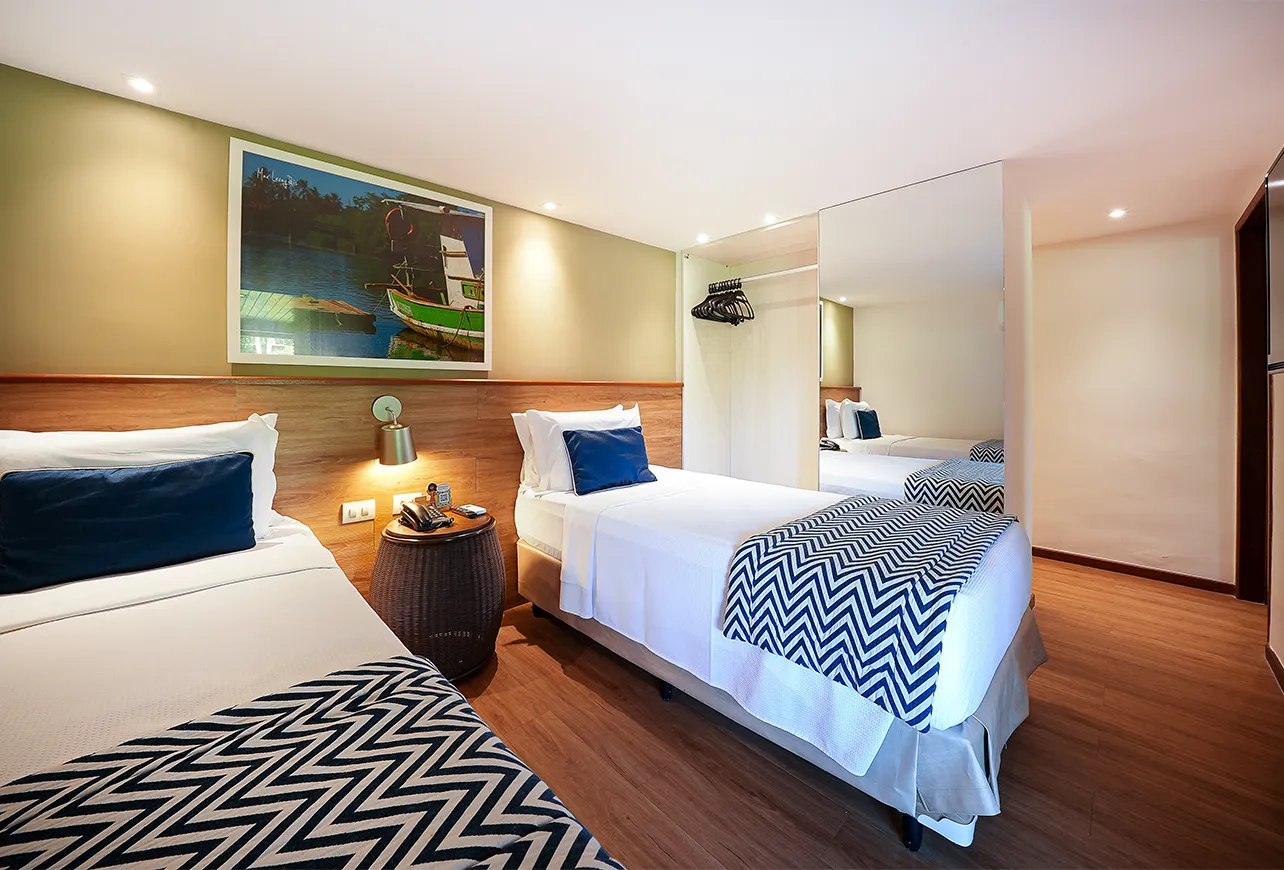 Segundo quarto da Suíte Familiar, com duas camas solteiro, uma mesinha de apoio e guarda-roupa entreaberto com espelho na porta, além de decoração em madeira e quadros.