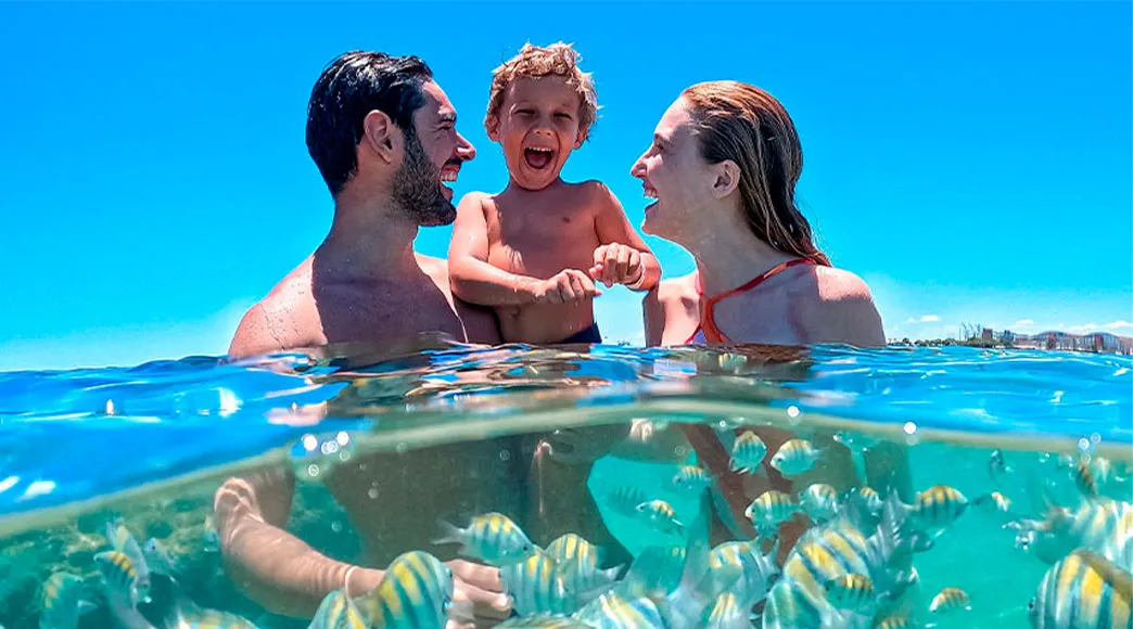 Foto dividida na linha d'água: na parte de cima, uma família feliz com um filho pequeno se diverte no mar; na parte de baixo, um cardume de peixes sargentinho nada ao redor deles na água cristalina.