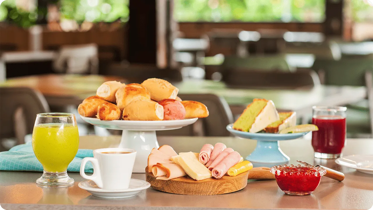 Mesa com pães, frios, queijos, bolos, geleia, café e sucos coloridos em ambiente iluminado