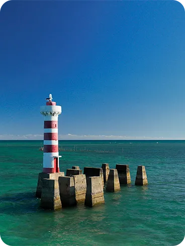 Farol listrado vermelho e branco sobre plataforma de concreto cercado por mar turquesa sob céu azul.