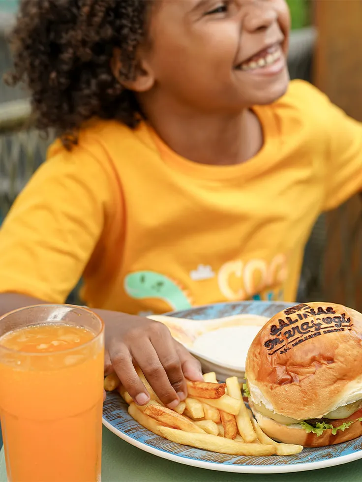 Criança comendo hambúrguer com logo do Salinas Maragogi, batatinha frita e um suco em área externa do resort.