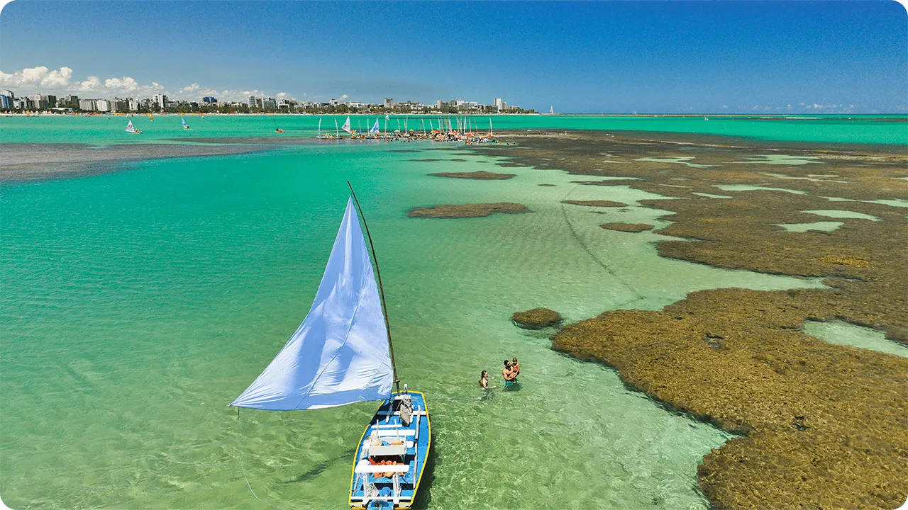 Vista aérea de las piscinas naturales de Pajuçara, Maceió, con una balsa de velas blancas en primer plano navegando por las aguas verde esmeralda. Al fondo, arrecifes de coral y la costa de la ciudad.