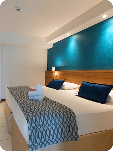 Cama queen size na acomodação Standard, em frente a roupeiro e painel de madeira com TV a cabo, mesa e cadeira.