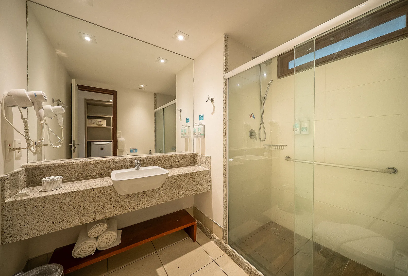 Banheiro da acomodação Standard, contendo área pra banho, instalações sanitárias, chuveiro com regulação de temperatura, decoração, amenities, toalhas, grande espelho, secador de cabelo e amenities pendurado na parede ao lado do espelho.
