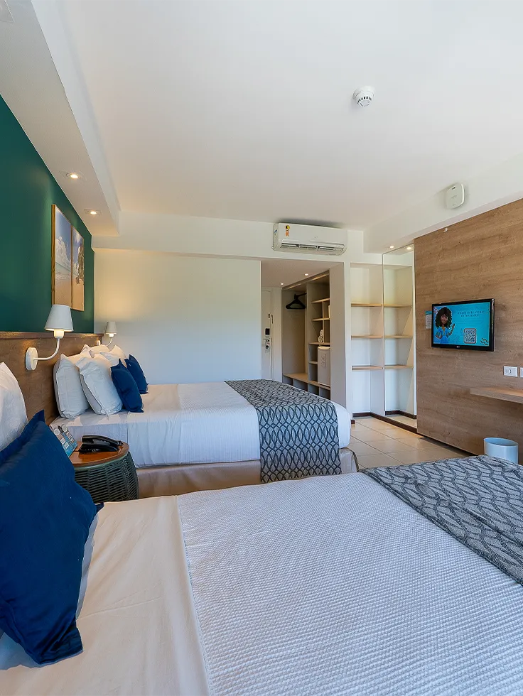 Vista lateral do quarto Apartamento Standard no Salinas Maceió com cama casal e cama solteiro. Em frente a cama de casal tem uma televisão pendurada ao lado da escrivaninha. Com decoração praiana com parede verde e quadros pendurados na parede.