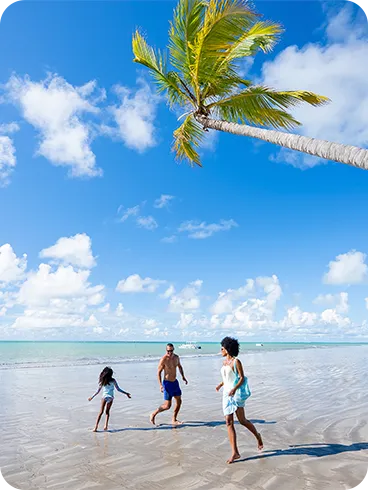 Una familia se divierte en la amplia playa de Ipioca en Maceió. Padre, madre e hija corren por la arena mojada bajo un cocotero inclinado, con el mar y el cielo azul de fondo.