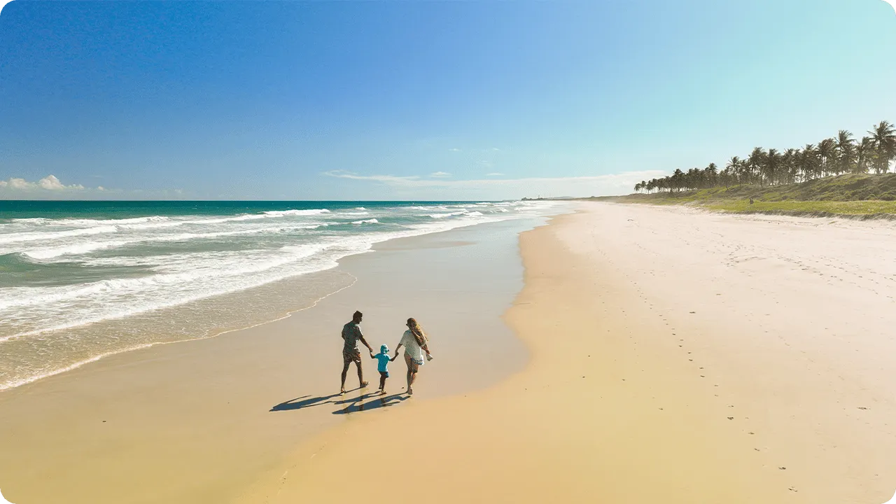 Grande faixa de areia clara da Praia do Francês com uma família caminhando de mãos dadas com mar azul-esverdeado ao fundo e linha de coqueiros à direita.