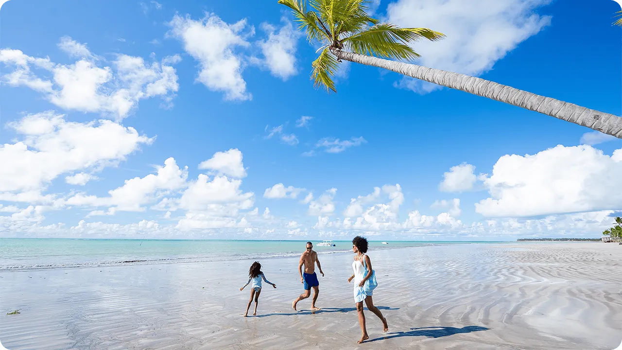 Una familia se divierte en la amplia playa de Ipioca en Maceió. Padre, madre e hija corren por la arena mojada bajo un cocotero inclinado, con el mar y el cielo azul de fondo.