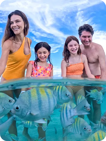 Fotografía sumergida y subacuática que muestra a una familia sonriente interactuando con bancos de peces de colores en aguas cristalinas, con los pies de la familia visibles en el fondo marino.