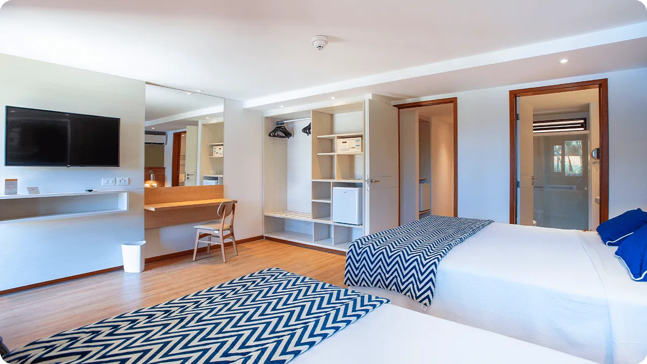 Una cama queen size y una cama individual en la Suite Familia, frente a una mesa y una silla, panel con TV por cable, armario, minibar, caja fuerte digital y baño.