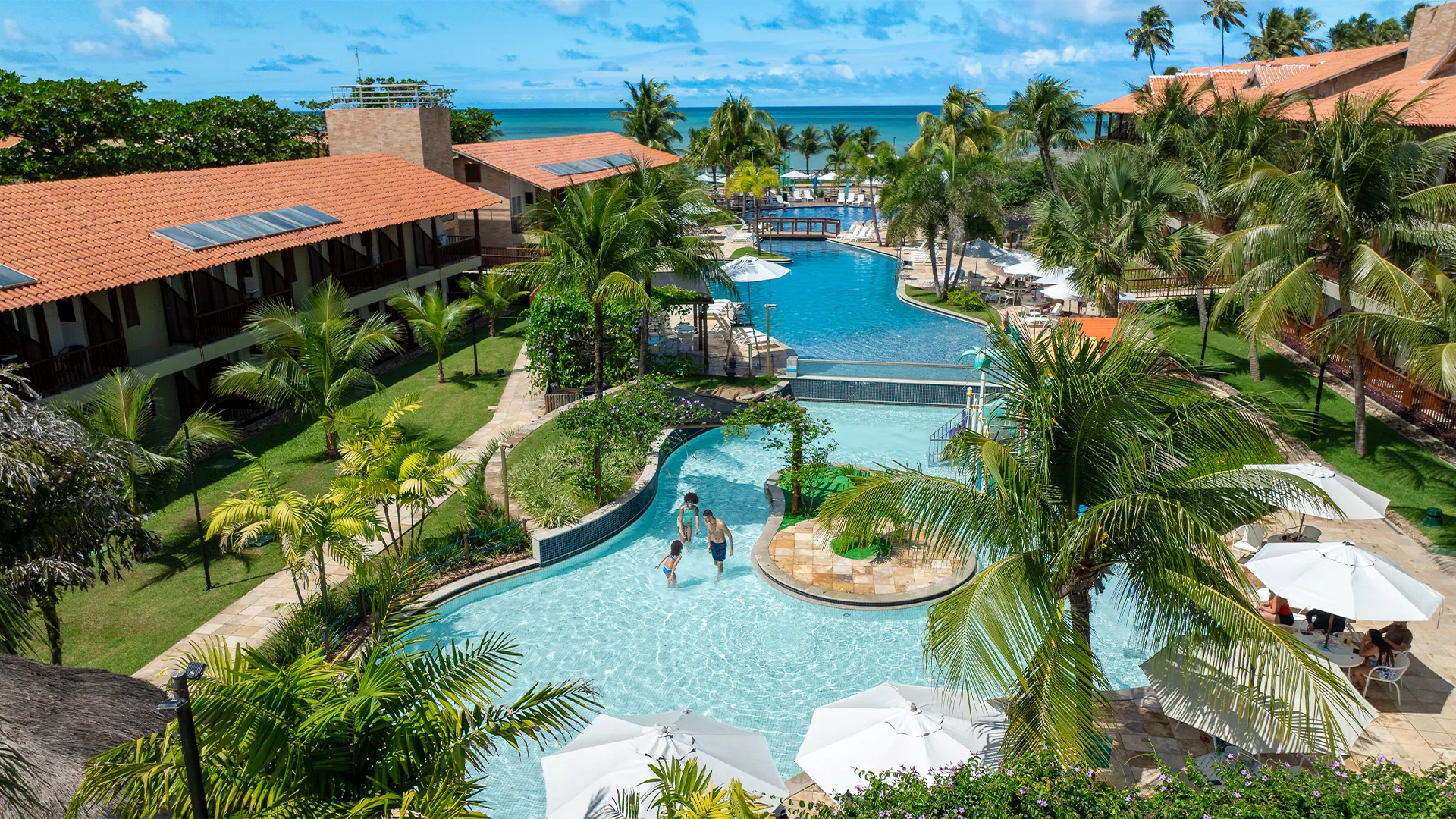 Vista aérea das piscinas e blocos de apartamentos do Salinas Maceió cercados por coqueirais sob céu azul. A família está na piscina infantil na área de lazer.