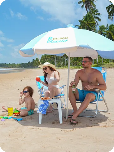 Família curtindo dia ensolarado na praia de Ipioca sob guarda-sol Salinas Maceió, com mar turquesa, areia dourada e uma vegetação magnífica ao fundo.