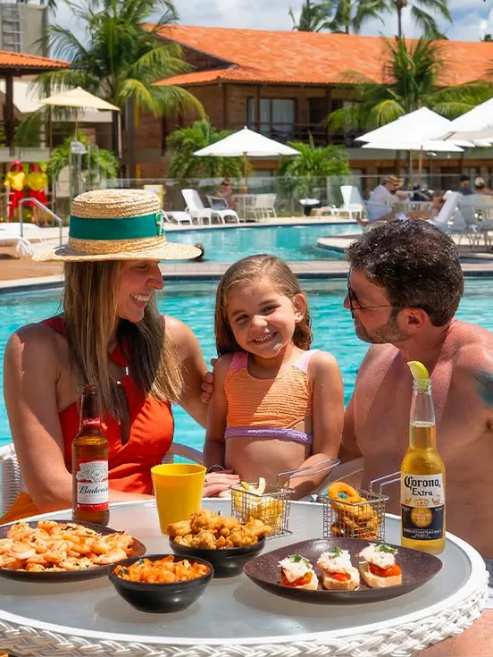 Família à beira da piscina do Salinas Maragogi saboreando camarões, Bruschettas outros petiscos e ao lado bebidas como a cerveja Budweiser e Corona Extra. Ao fundo piscina, espreguiçadeiras, guarda-sóis e estrutura de acomodações.