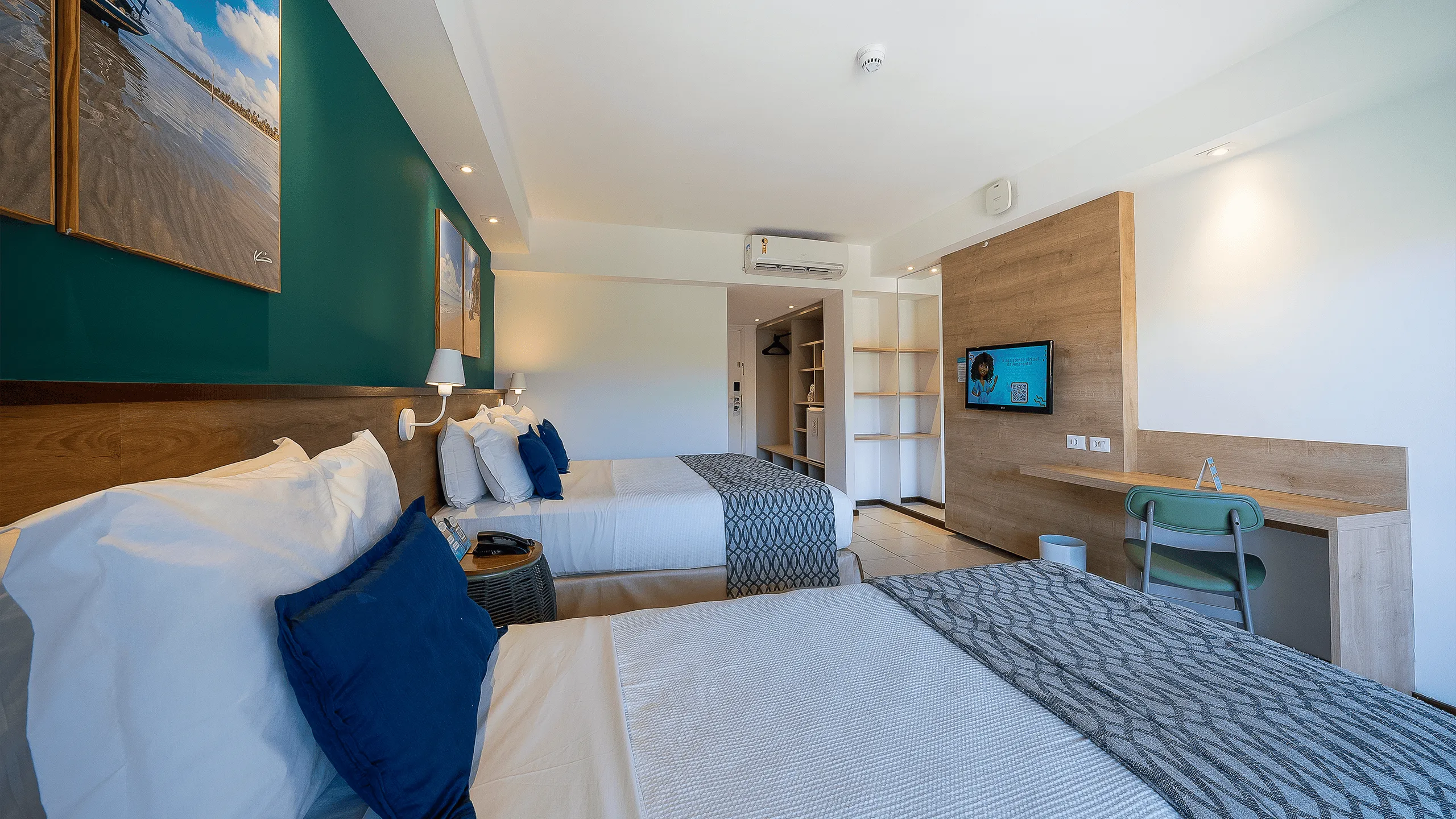 Vista lateral do quarto Apartamento Standard no Salinas Maceió com cama casal e cama solteiro. Em frente a cama de casal tem uma televisão pendurada ao lado da escrivaninha. Com decoração praiana com parede verde e quadros pendurados na parede.