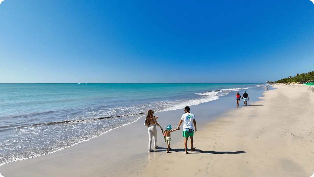 Famílias caminhando de mãos dadas pela areia molhada na Praia de Jatiúca, com mar calmo e céu azul.