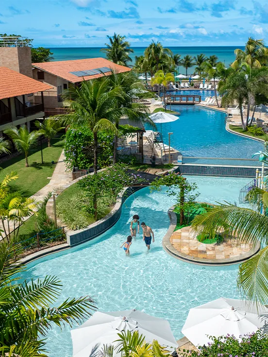 Vista aérea das piscinas e blocos de apartamentos do Salinas Maceió cercados por coqueirais sob céu azul. A família está na piscina infantil na área de lazer.