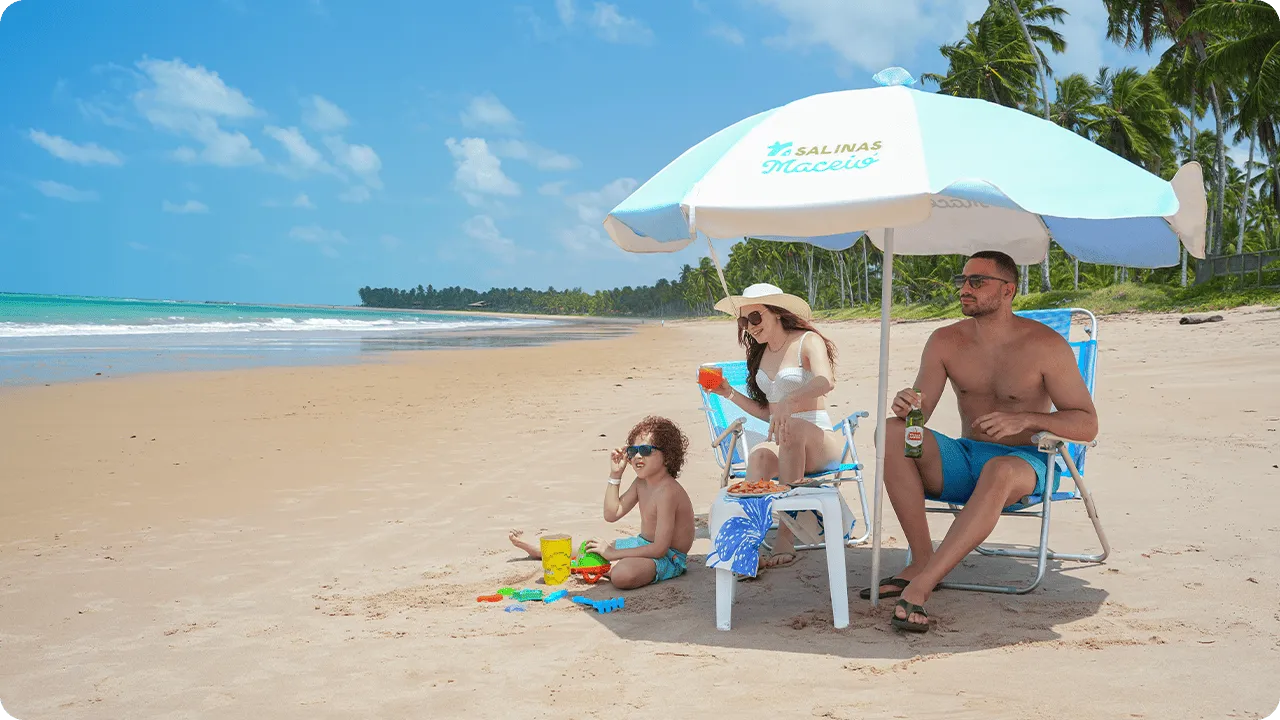 Família curtindo dia ensolarado na praia de Ipioca sob guarda-sol Salinas Maceió, com mar turquesa, areia dourada e uma vegetação magnífica ao fundo.