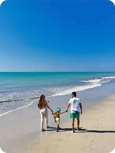 Famílias caminhando de mãos dadas pela areia molhada na Praia de Jatiúca, com mar calmo e céu azul.