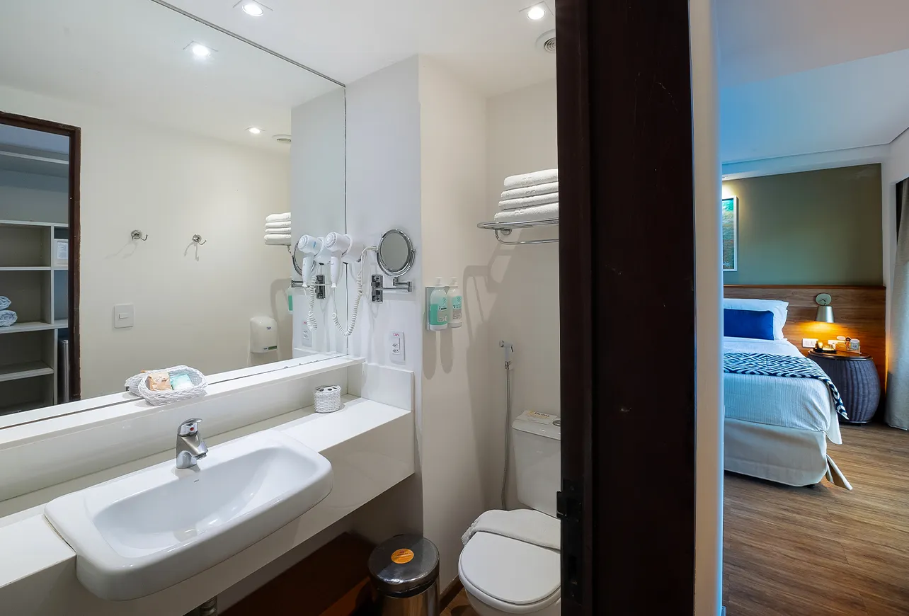 Banheiro da acomodação Praia Frente Mar, com áreas sanitárias, amenities O Boticário, toalhas e secador de cabelo. Pelo espelho, é possível ver o roupeiro com toalhas dentro e espaços pra guardar as roupas.