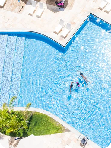 Vista aérea de la piscina de Salinas Maceió, con tres personas nadando en sus aguas cristalinas. Alrededor de la piscina hay una terraza de piedra con varias tumbonas blancas y cocoteros.