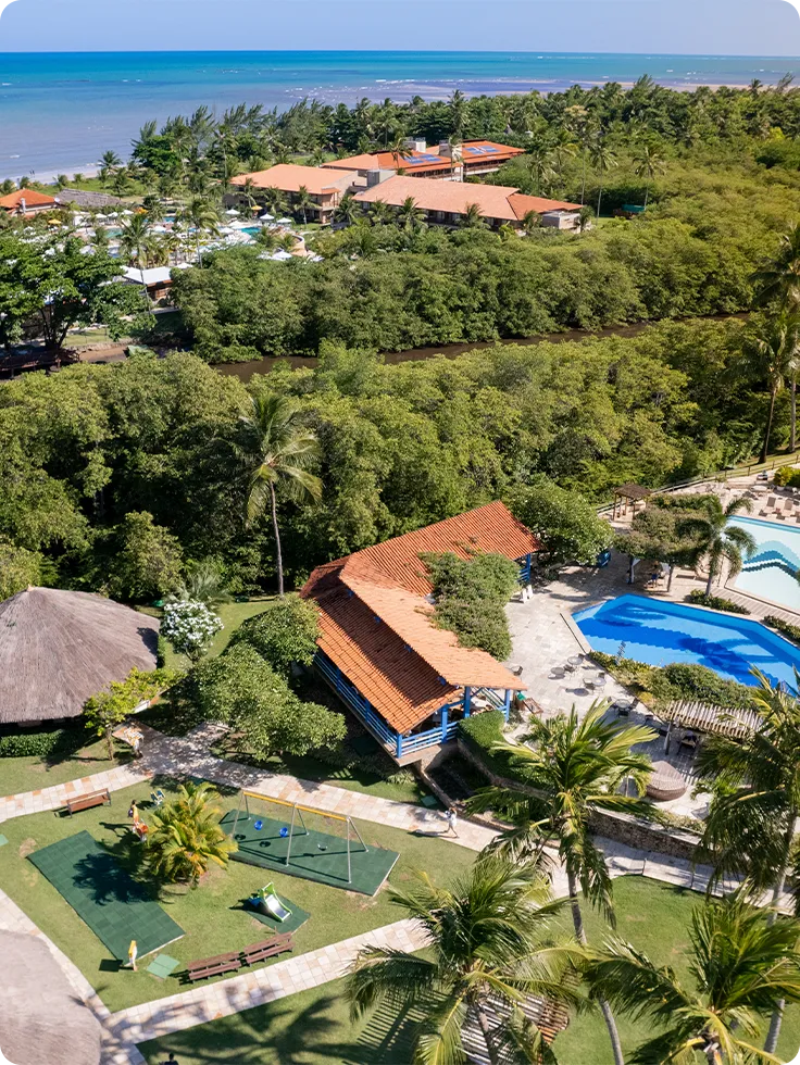 Vista aérea do Salinas Maragogi cercada por vegetação bem verde. Com piscinas superiores, o Café Canoas e o teen space. Ao fundo se ver um pouco a área das piscinas, o mar bem azul e o céu sem nuvens.