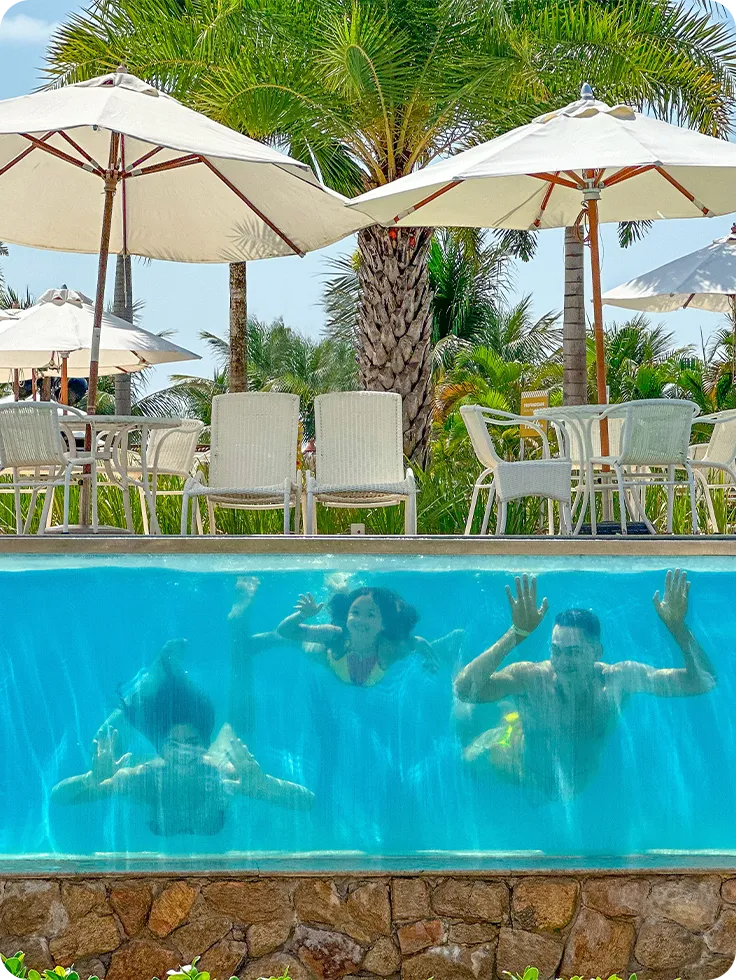 Família mergulhando e sorrindo debaixo d'água, em piscina com parede de vidro no Salinas Maragogi, acima tem guarda-sóis brancas com palmeira ao fundo.