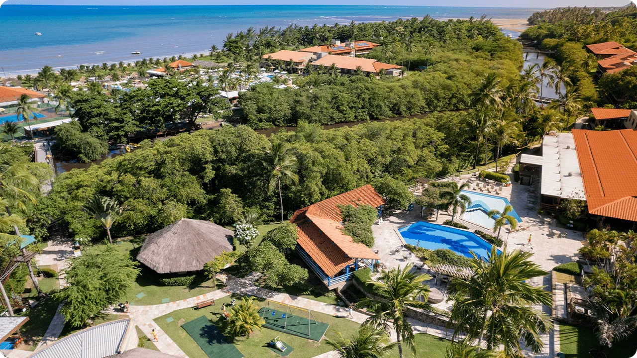 Vista aérea do Salinas Maragogi cercada por vegetação bem verde. Com piscinas superiores, o Café Canoas e o teen space. Ao fundo se ver um pouco a área das piscinas, o mar bem azul e o céu sem nuvens.