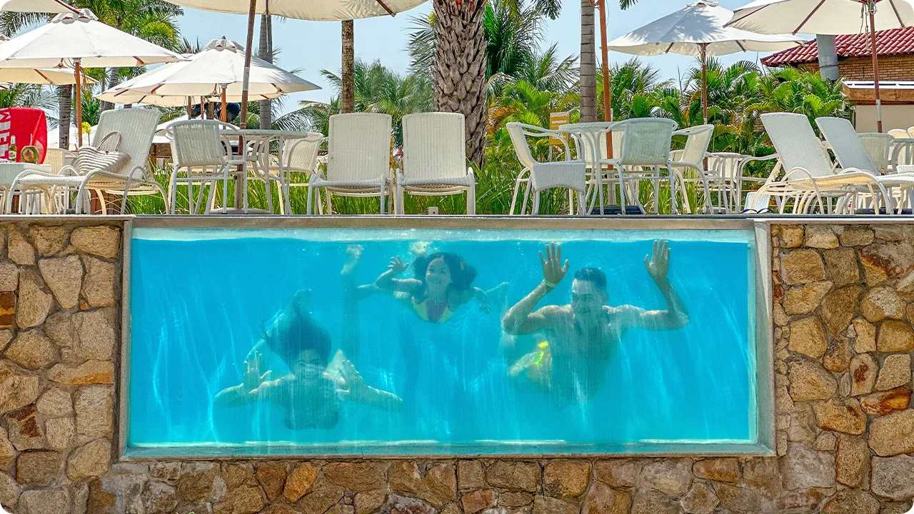 Família mergulhando e sorrindo debaixo d'água, em piscina com parede de vidro no Salinas Maragogi, acima tem guarda-sóis brancas com palmeira ao fundo.