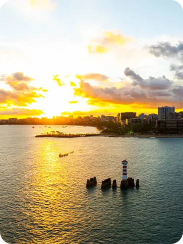 Vista da Praia de Ponta Verde ao pôr do sol alaranjado com farol cercado por pedras, mar calmo, barcos distantes e prédios iluminados.