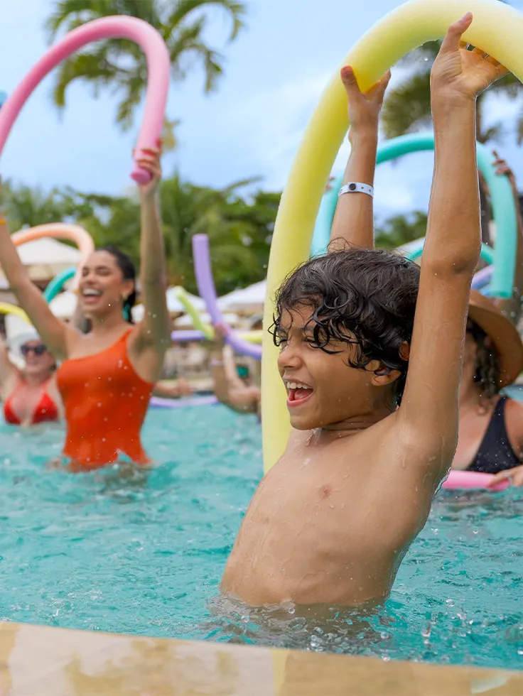 Hóspedes com boias praticando atividade física em piscina do Salinas Maragogi, tem guarda-sóis brancas com palmeira ao fundo.