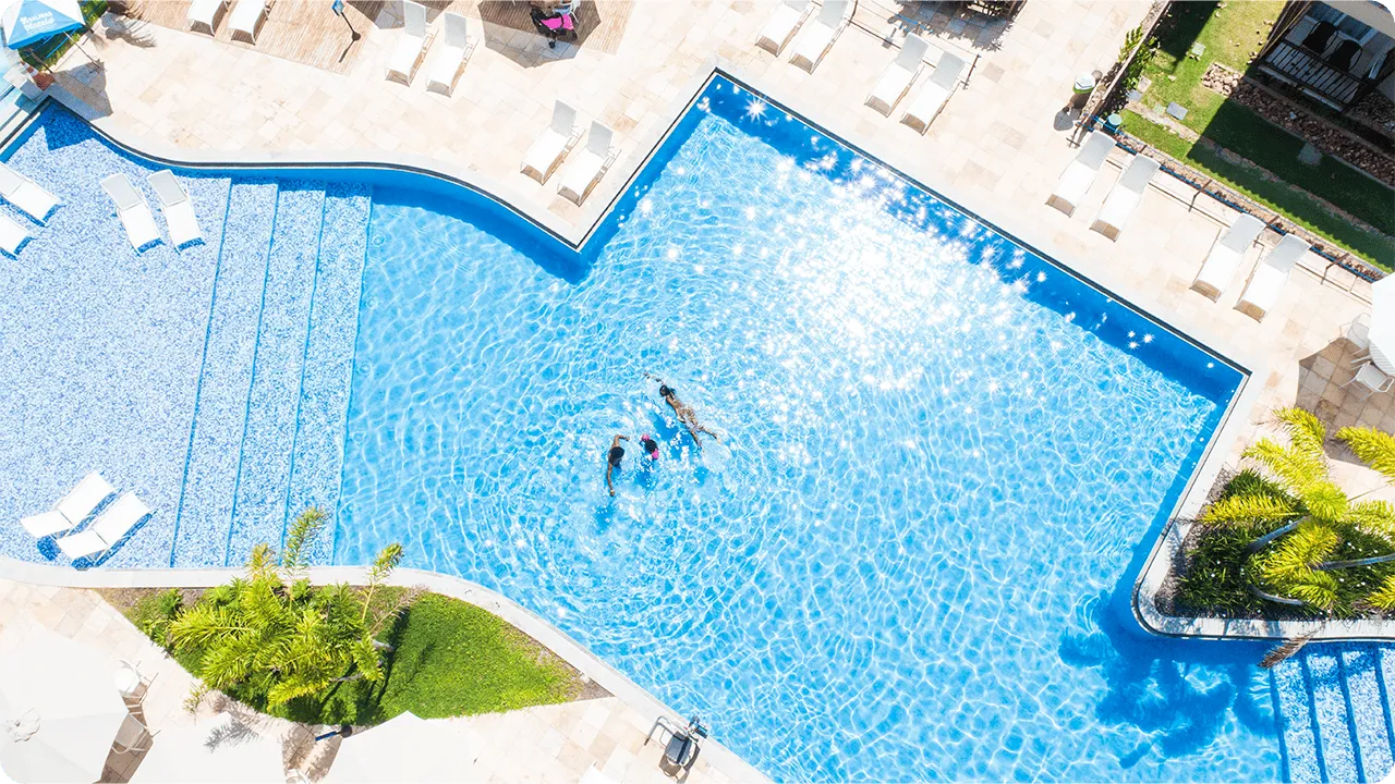 Vista aérea de la piscina de Salinas Maceió, con tres personas nadando en sus aguas cristalinas. Alrededor de la piscina hay una terraza de piedra con varias tumbonas blancas y cocoteros.