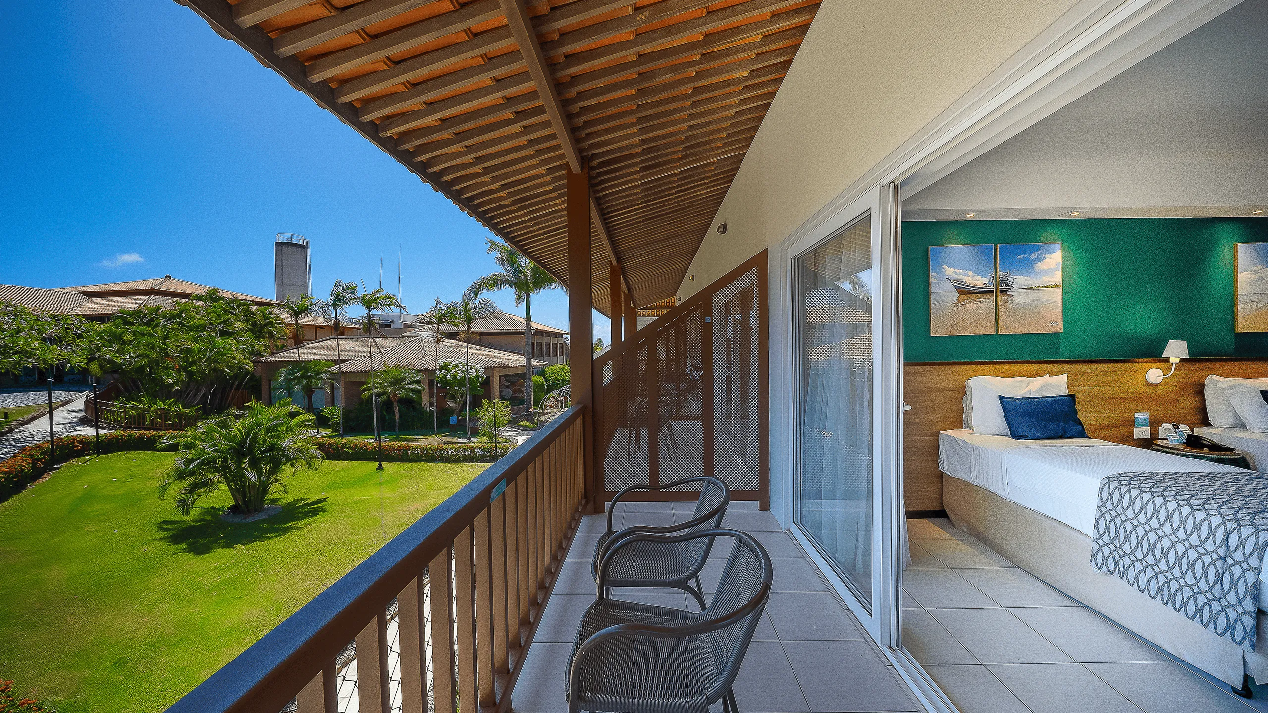 Varanda do Apartamento Standard no Salinas Maceió, ao lado esquerdo a vista pra jardins e área verde do resort e ao lado direito a parte interna do quarto com cama de solteiro.