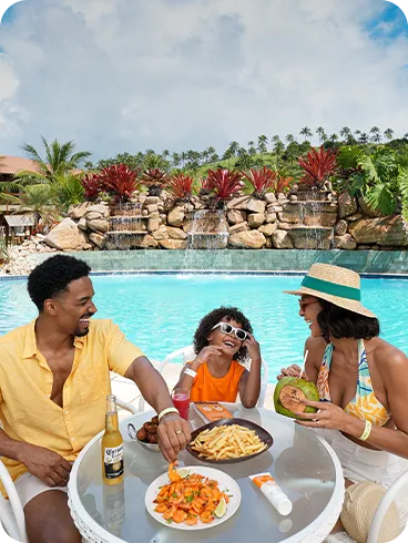 Família sentada à mesa na área da piscina do Salinas Maragogi, com cascata e jardim ao fundo; na mesa, camarões ao molho, batata frita, bolinhos e bebidas como cerveja, suco e água de coco.