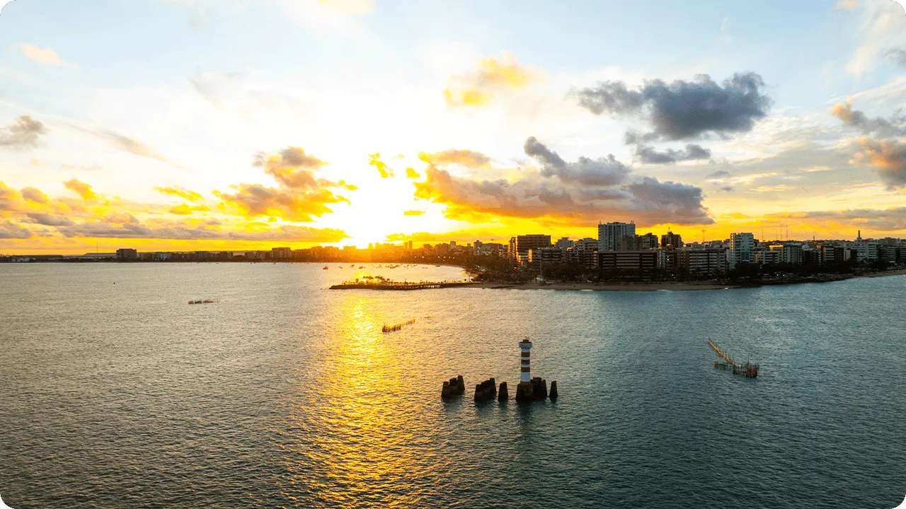 Vista da Praia de Ponta Verde ao pôr do sol alaranjado com farol cercado por pedras, mar calmo, barcos distantes e prédios iluminados.