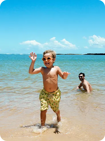 Menino com óculos de sol correndo animado pela beira da praia enquanto um adulto se diverte na água ao fundo