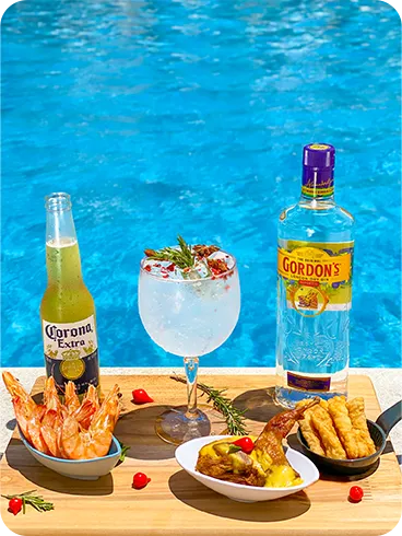 Taça de gin tônica com especiarias, garrafa de Stella Artois e petiscos sobre tábua de madeira junto à piscina