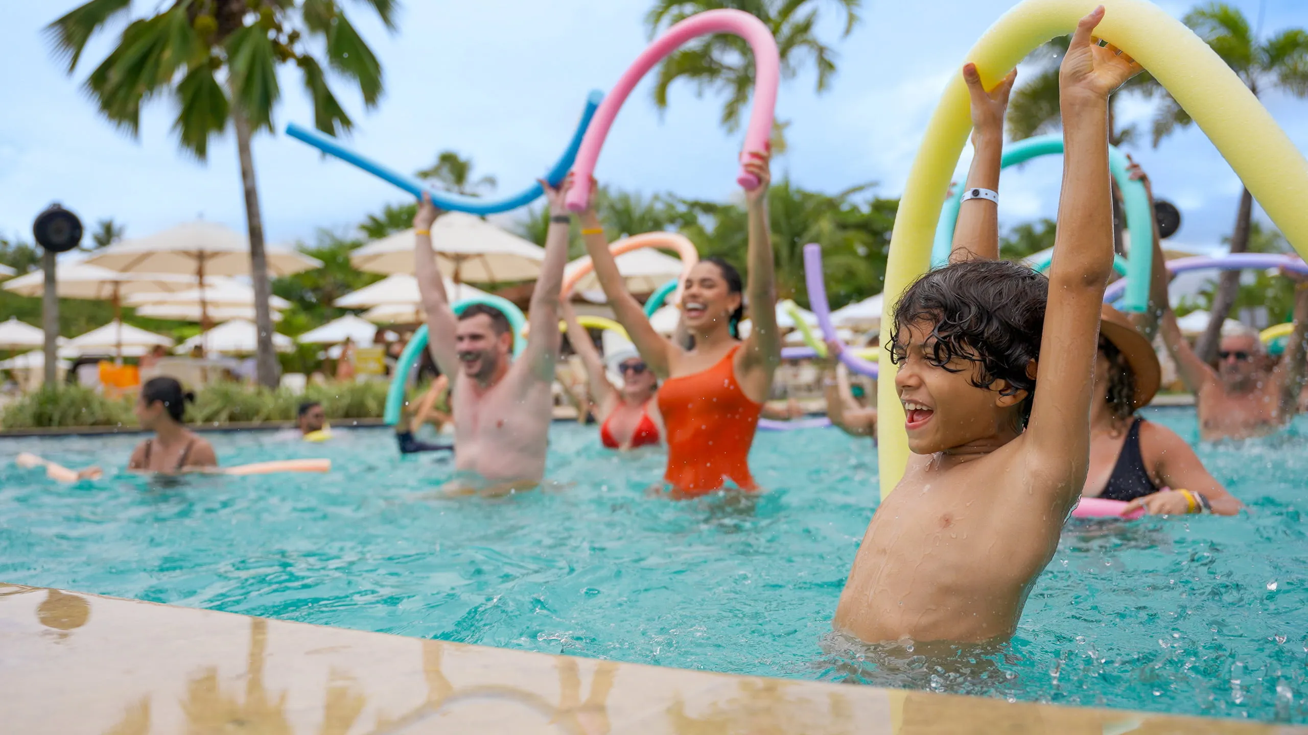 Hóspedes com boias praticando atividade física em piscina do Salinas Maragogi, tem guarda-sóis brancas com palmeira ao fundo.