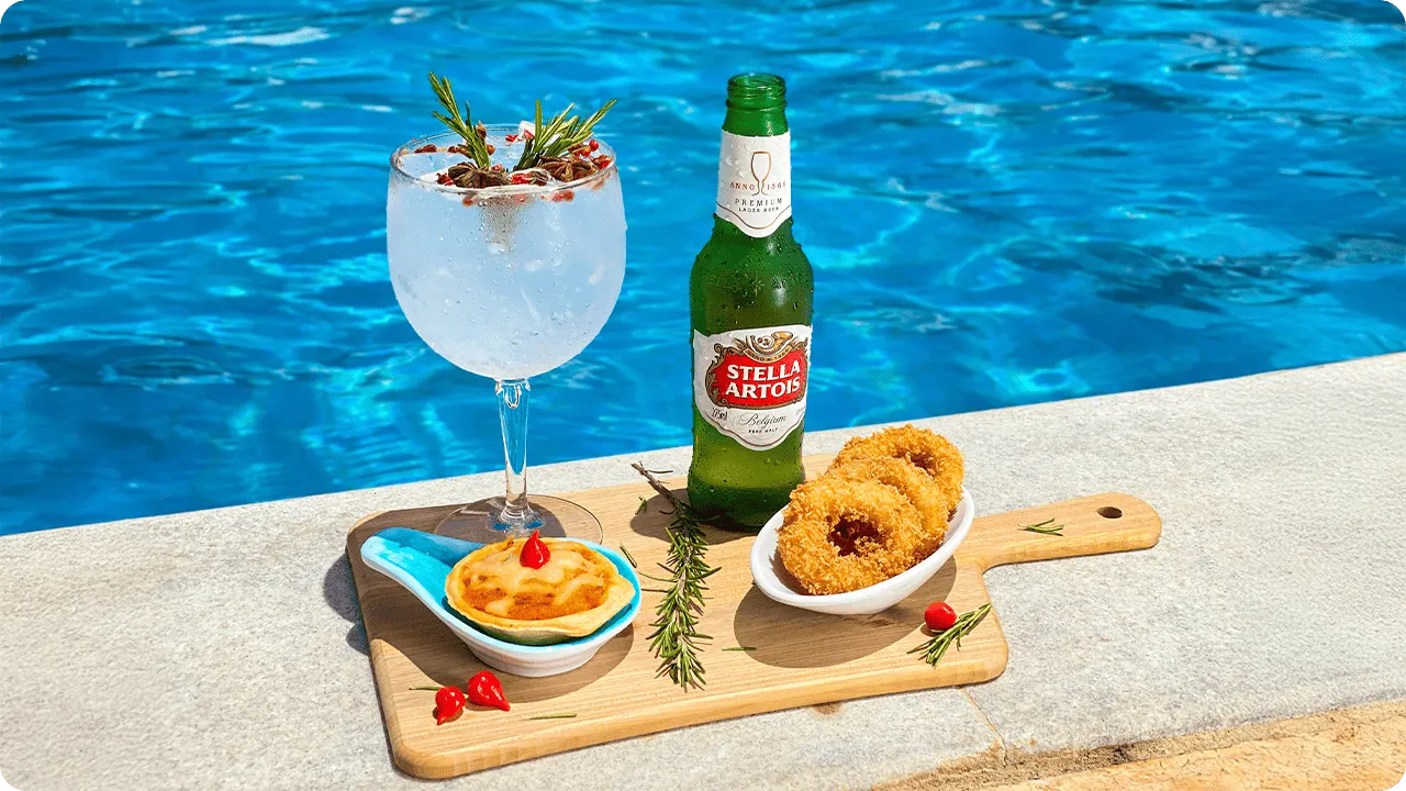 Taça de gin tônica com especiarias, garrafa de Stella Artois e petiscos sobre tábua de madeira junto à piscina