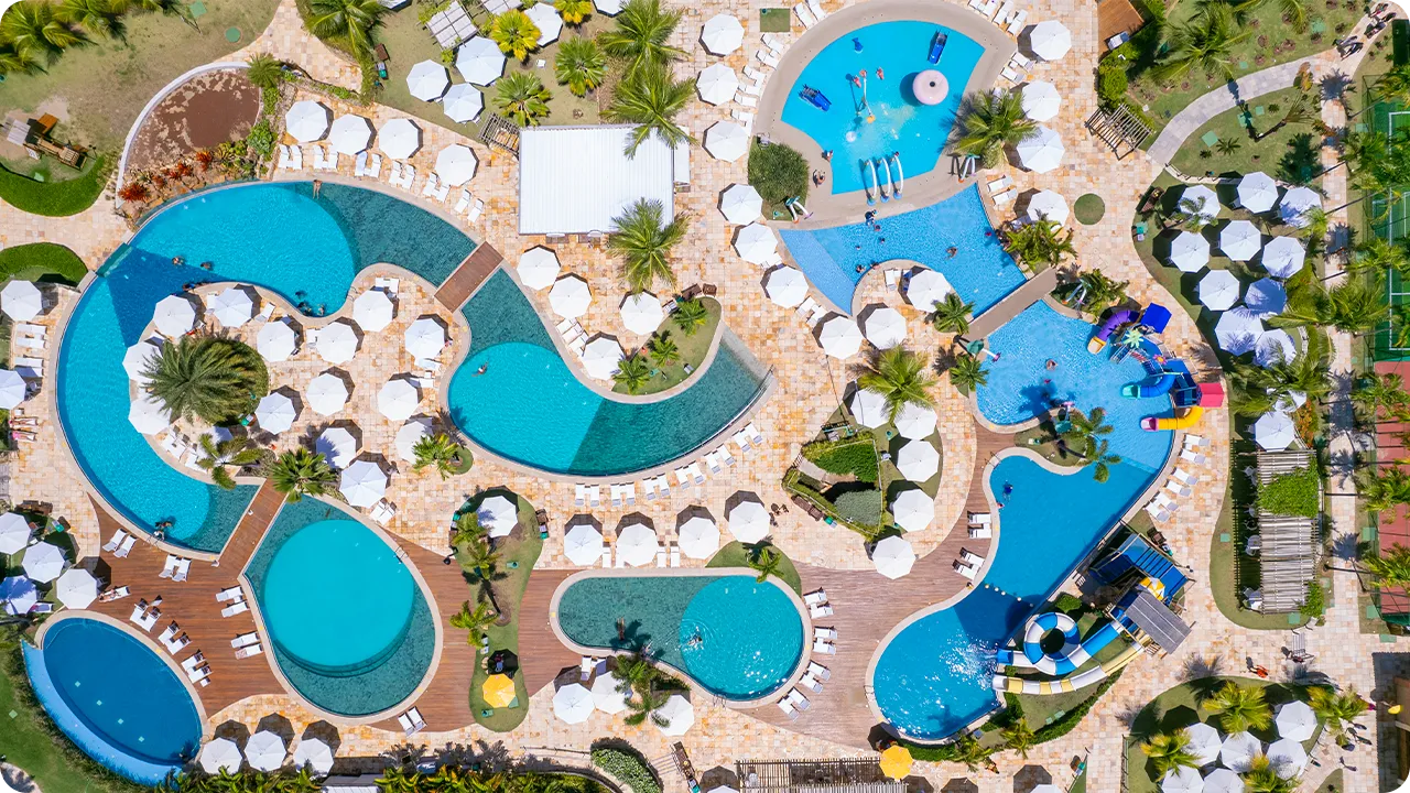 Vista aérea del gran complejo de piscinas de Salinas Maragogi, que muestra las piscinas de formas orgánicas, las terrazas de madera, las sombrillas blancas y el parque acuático infantil.