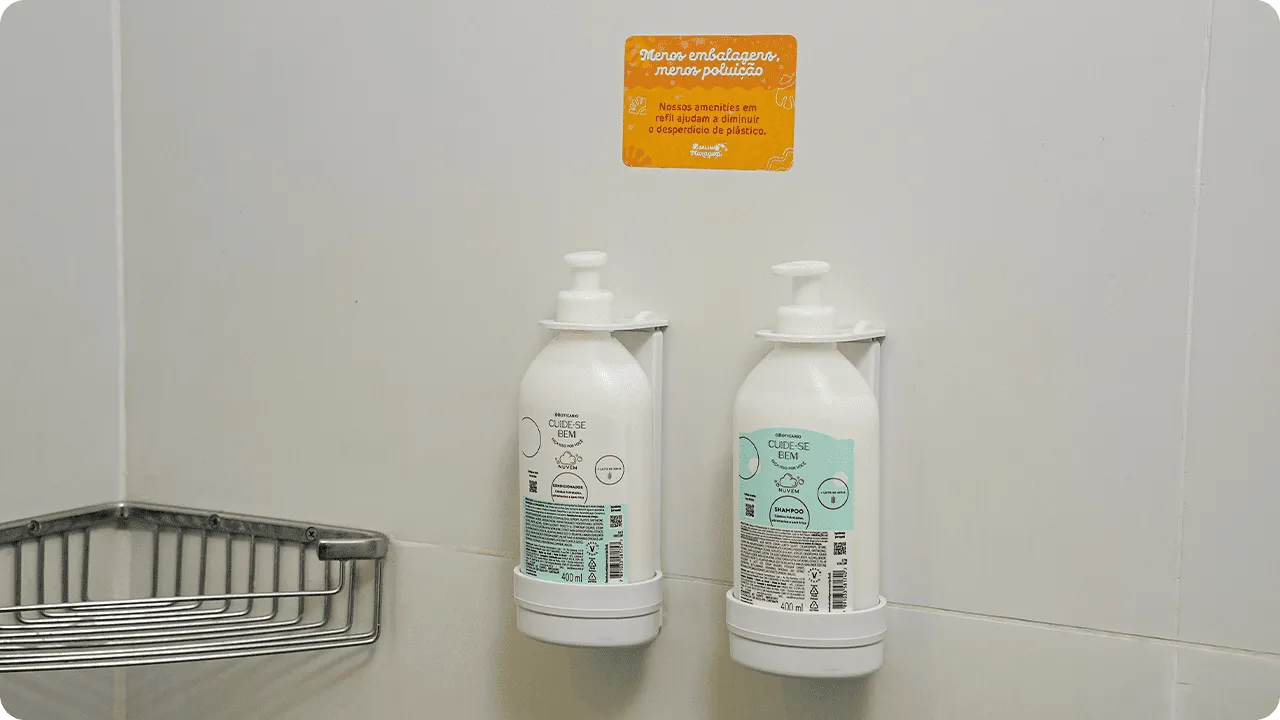 Shampoo e condicionador O Boticário em dispensers de refil na parede do banheiro, acompanhados de um aviso sobre sustentabilidade e redução de plástico.