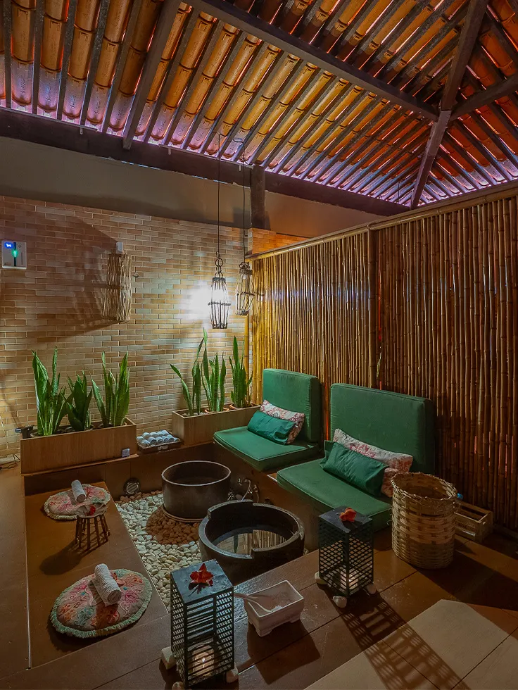 Espaço de SPA com decoração aconchegante e rústica, poofs verdes em cima do banco de madeira, cestas com toalhas enroladas, iluminação suave com velas e luminárias pendentes, parede de bambu e plantas decorativas.