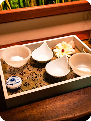 Decorative tray on a rustic wooden table with white ceramic pots, artificial flower, decorative soap, mini white iron chair and amber vase with lilac flowers.